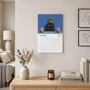 May include: A wall calendar for November 2026 featuring an illustration of a black cat with a green frog on its head under a red mushroom cap, flanked by two bubble tea drinks against a blue background, with a grid of dates below the image.