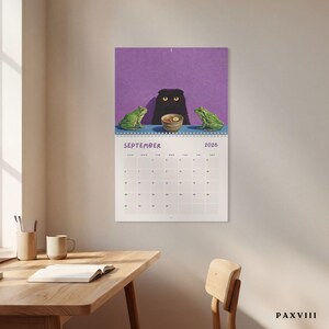 May include: A wall calendar for September 2026 featuring an illustration of a black cat with yellow eyes sitting at a blue table between two green frogs. A small bowl of food sits in front of the cat. The background is a solid purple color.