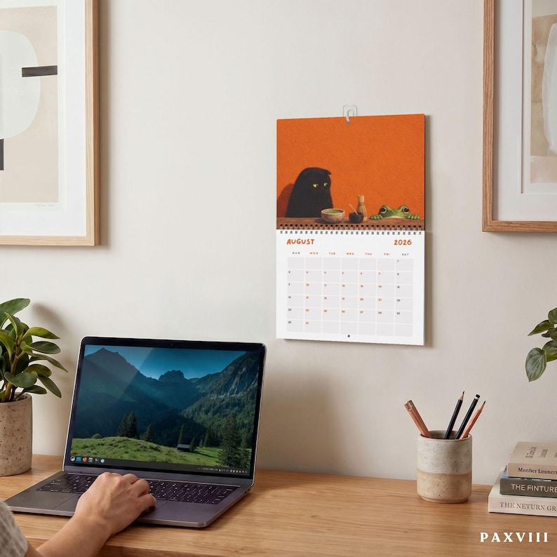 May include: A wall calendar for August 2026 featuring an illustration of a black cat and a green frog sitting at a table against an orange background. The calendar grid is white with black text and orange numbers, hanging on a light wall.