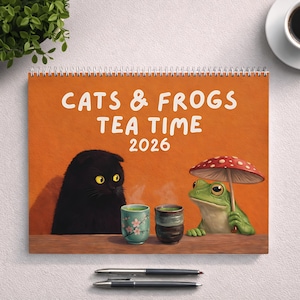 May include: A spiral-bound calendar titled "CATS & FROGS TEA TIME 2026" features a black cat and a green frog under a mushroom umbrella. Two teacups sit on a wooden surface, with two pens below. The background is orange.