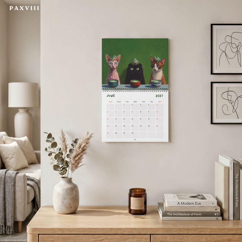 May include: A wall calendar for June 2021 featuring an illustration of three cats sitting at a table with small bowls and frogs on their heads. The calendar has a green background, a white grid for dates, and is displayed on a light wall.
