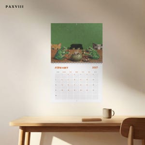 May include: A wall calendar for February 2027 featuring an illustration of two green frogs sitting at a table with a teapot and snacks, observed by three cats peering over the edge. The text PAXVIII is printed in the top left corner of the image.