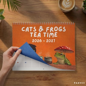 May include: A spiral-bound wall calendar with an orange cover featuring the text CATS & FROGS TEA TIME 2026 - 2027. The illustration shows a green frog holding a red and white spotted mushroom umbrella next to a steaming tea cup.