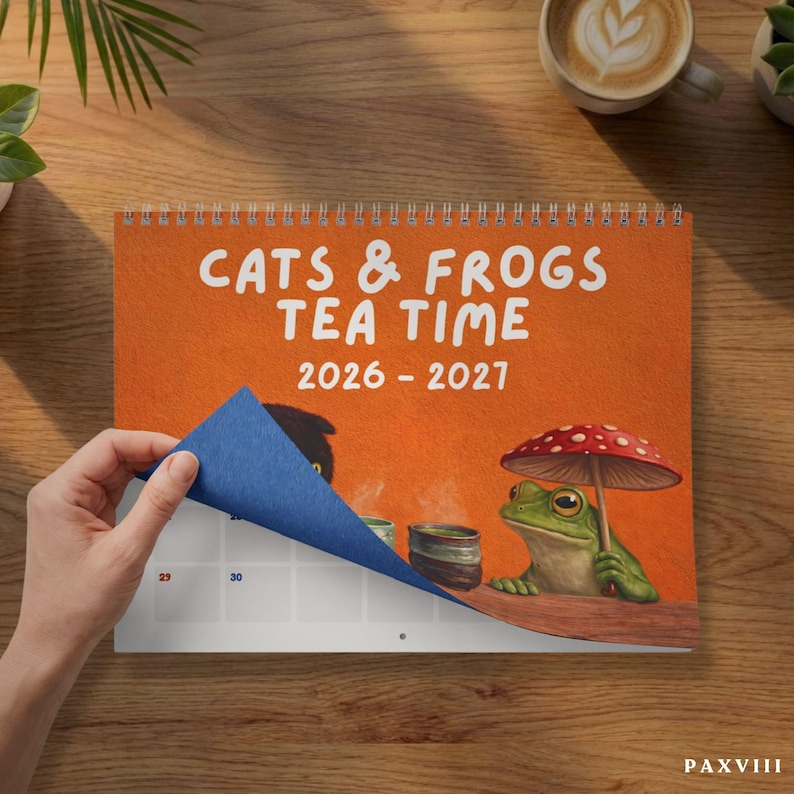 May include: A spiral-bound wall calendar with an orange cover featuring the text CATS & FROGS TEA TIME 2026 - 2027. The illustration shows a green frog holding a red and white spotted mushroom umbrella next to a steaming tea cup.