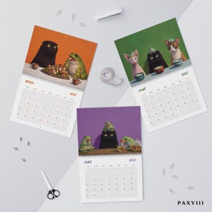 May include: Three wall calendar pages for April, May, and June 2027 featuring illustrations of cats and frogs. The April page shows a black cat and a frog with mushrooms. The May page shows a black cat with frogs. The June page shows three cats.