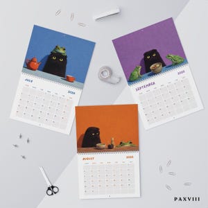 May include: Three 2026 wall calendar pages for July, August, and September. Each page features a black cat and green frogs in various settings with tea sets. The July page is blue, August is orange, and September is purple. Includes paper clips and scissors.