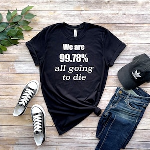 May include: Black t-shirt with white text that reads "We are 99.78% all going to die". The shirt is laid flat on a woodgrain surface with a pair of black and white sneakers, blue jeans, and a black baseball cap with a white Adidas logo.