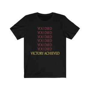 Op de afbeelding: Zwart T-shirt met de tekst "YOU DIED" zes keer herhaald in roze en "VICTORY ACHIEVED" in geel.