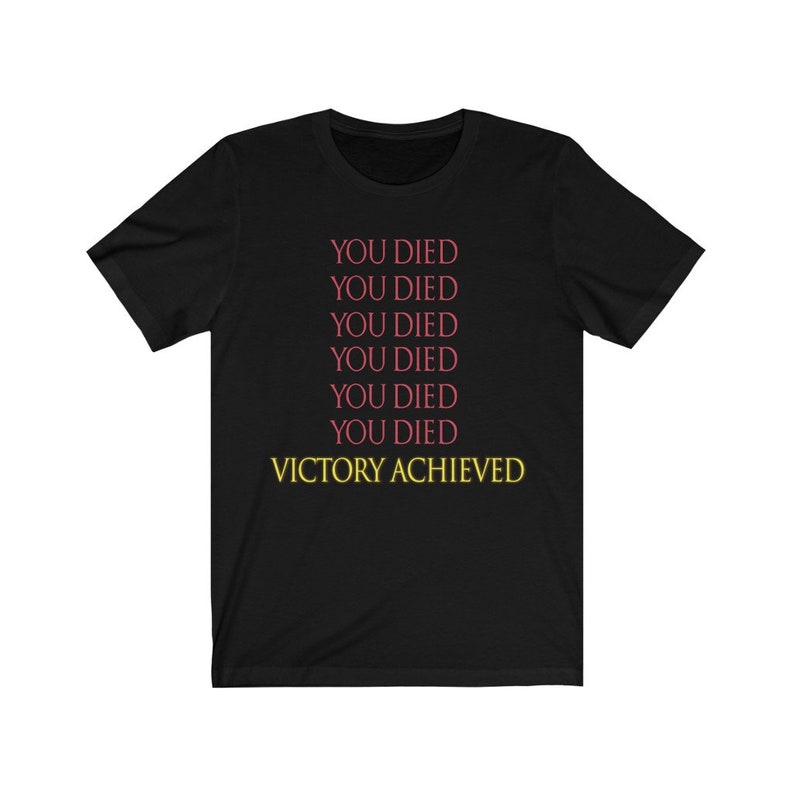 Op de afbeelding: Zwart T-shirt met de tekst "YOU DIED" zes keer herhaald in roze en "VICTORY ACHIEVED" in geel.