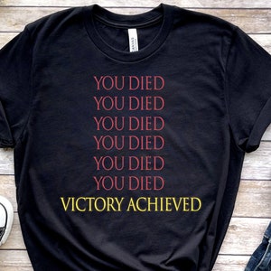 Op de afbeelding: Zwart T-shirt met de tekst "YOU DIED" zes keer herhaald in rood, gevolgd door "VICTORY ACHIEVED" in geel.