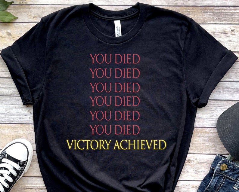 Op de afbeelding: Zwart T-shirt met de tekst "YOU DIED" zes keer herhaald in rood, gevolgd door "VICTORY ACHIEVED" in geel.