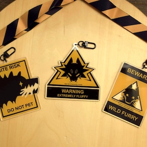 Puede incluir: Tres señales de advertencia con diseños y texto en negro y amarillo. Una señal dice "BITE RISK DO NOT PET" con una silueta de lobo. Otra dice "WARNING EXTREMELY FLUFFY". La tercera dice "BEWARE! WILD FURRY". También hay un cordón a rayas negro y dorado.