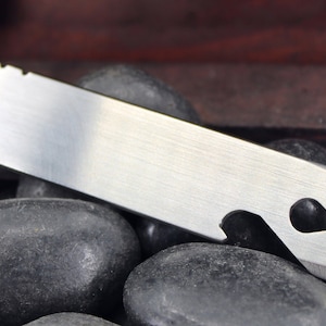 May include: A silver multi-tool with the word "Wrencher" engraved on it. The tool is in a wooden box with black stones.