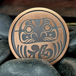 May include: A round, copper-colored coin featuring a Daruma doll design. The coin has a dark, etched Daruma face with the Japanese character for "fortune" at the bottom. The coin is resting on dark, smooth stones.