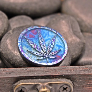 Op de afbeelding: Een ronde, blauwe, paarse en groene metalen munt met een marihuanabladontwerp.