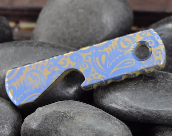 May include: A blue and gold patterned titanium bottle opener with a hole for a keychain. The opener is sitting on a bed of smooth, grey stones.
