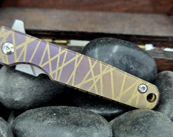 Könnte beinhalten: Ein kleines Taschenmesser mit einem purpurfarbenen und goldenen Griff. Das Messer hat eine teilweise geöffnete Klinge und eine kleine Schraube am Griff.