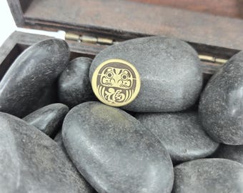 May include: A gold-colored metal charm with a detailed design of a Japanese Daruma doll. The charm is sitting on a bed of smooth, gray stones.