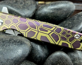 Könnte beinhalten: Ein Taschenmesser mit einem lila-goldenen Wabenmuster. Das Messer liegt auf dunklen grauen Steinen. Die Klinge ist silberfarben und teilweise geöffnet. Die Länge des Messers beträgt ca. 10 cm.