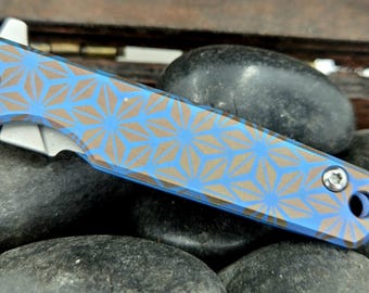 Könnte beinhalten: Ein blaues und braunes Taschenmesser mit einem geometrischen Muster auf dem Griff. Das Messer ist geschlossen und liegt auf einem Bett aus glatten, grauen Steinen.