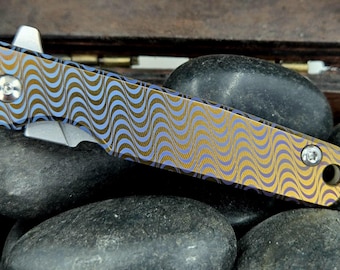 Könnte beinhalten: Ein kleines Taschenmesser mit einem wellenförmigen Muster in Blau und Gold auf dem Griff. Die Klinge ist teilweise sichtbar und hat eine silberne Oberfläche.