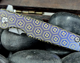 Könnte beinhalten: Ein Klappmesser mit gemustertem Griff. Der Griff hat ein blau-goldenes sechseckiges Design mit floralen Akzenten. Die Klinge ist silberfarben und das Messer liegt auf schwarzen Steinen.