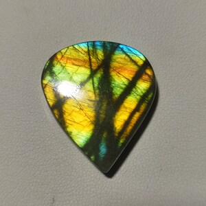 Pode incluir: Um cabochão de labradorita em forma de lágrima com um brilho iridescente verde, amarelo e azul.