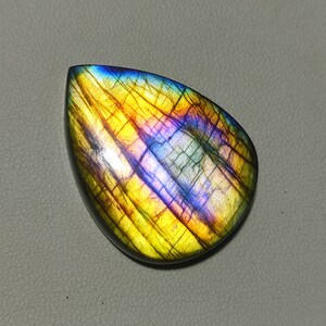 Pode incluir: Um cabochão de labradorita em forma de lágrima com cores iridescentes azuis, amarelas e laranja.