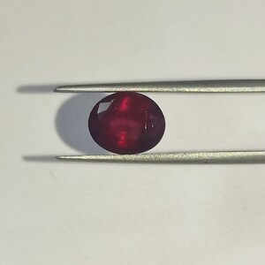May include: A deep red gemstone with a faceted cut, held in tweezers against a white background.