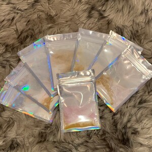 May include: Six clear plastic resealable bags with iridescent holographic foil accents, each containing a different colored powder. The powders are various shades of brown, pink, and purple.