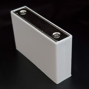 Puede incluir: Una batería rectangular blanca con dos tornillos visibles en la parte superior.