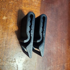 May include: Two black 3D-printed plastic clips with a U-shaped profile. Each clip features a serrated inner edge designed for gripping, a curved top section, and a flat base with a small protruding tab, showing visible layer lines from the printing process.