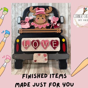 以下が含まれることがあります： ピンクの帽子をかぶり、バレンタインデーのハートを持ったヘラジカが描かれた、黒いトラック型の木製装飾。トラックの荷台には、ハート型の文字で「LOVE」と書かれています。「FINISHED ITEMS MADE JUST FOR YOU」の文字が下部にあります。