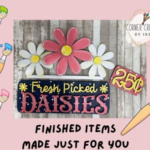 May include: Wooden sign featuring three daisies, a price tag, and paintbrushes. The sign displays "Fresh Picked Daisies" in pink and yellow lettering on a navy blue background. The price tag reads "25p".