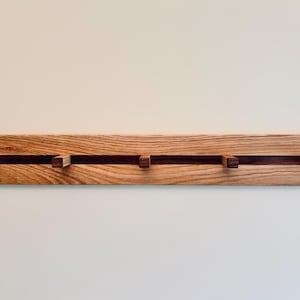 May include: Wooden wall-mounted coat rack with four hooks. The rack is made of light-coloured wood with a dark wood accent running horizontally across the centre. The hooks are rectangular and made of the same dark wood.