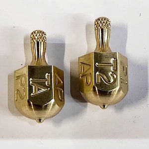 May include: Two brass dreidels, each with a textured top and a hexagonal body. The sides of the dreidels are engraved with Hebrew letters. The dreidels are displayed on a white surface.