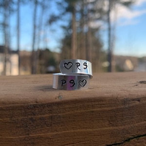May include: A silver ring with the initials "PS" and "RS" engraved on it, separated by a heart symbol on each side.