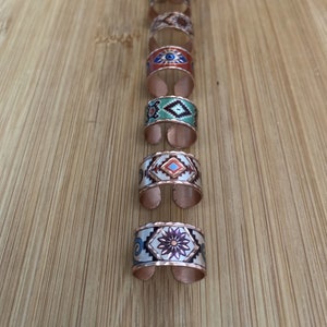Peut inclure: Une collection de bagues réglables de couleur cuivre avec des motifs émaillés colorés. Les bagues présentent divers motifs, notamment des formes géométriques, des motifs floraux et un symbole d'œil. Les bagues sont présentées sur une surface en bois.