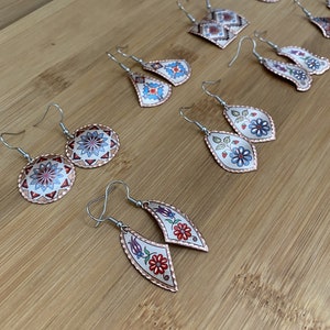 Könnte beinhalten: Eine Sammlung handgefertigter Kupferohrringe mit verschiedenen floralen und geometrischen Designs. Die Ohrringe sind in leuchtenden Farben bemalt und haben einen weißen Hintergrund.
