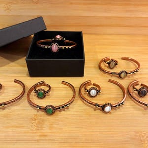 May include: Copper-coloured bracelets and rings with assorted gemstone embellishments. The jewellery is arranged on a wooden surface, with one set in a black box. Gemstones include turquoise, green, white, and pink stones.