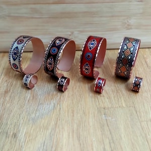 May include: Four copper bracelets with different colourful patterns, including an eye design, geometric shapes, and a white and blue design. Each bracelet has a matching ring.