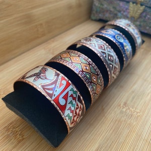 Puede incluir: Cinco pulseras de cobre con patrones geométricos coloridos y un acabado de cobre. Las pulseras están apiladas en un expositor de terciopelo negro.
