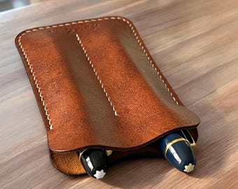 May include: A brown leather pen case with white stitching, holding two pens. One pen is black, the other is blue with gold accents. The case is on a wooden surface.