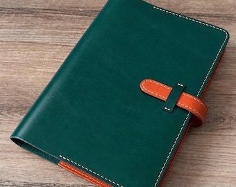 Puede incluir: Una funda de cuero para cuaderno en color verde oscuro con acabado liso y costuras blancas en contraste. La funda incluye un cierre de correa de cuero naranja que asegura la solapa frontal, con bordes de cuero naranja visibles.
