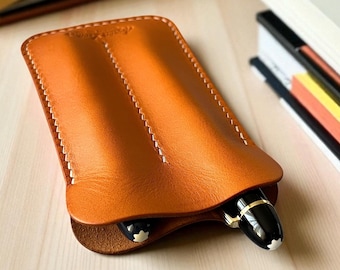 May include: A handcrafted, orange-brown leather pen case with white stitching. Two black and gold pens are partially visible inside the case. The case is on a light wood surface, with books in the background.