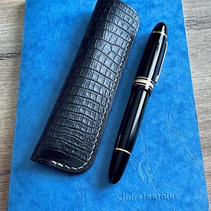 Op de afbeelding: Een zwarte pen met gouden accenten en een zwart leren pennenetui op een blauw notitieboek met de tekst "Clairefontaine" in reliëf op de omslag.