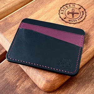 Può includere: Un porta carte in pelle nera con un accento in pelle bordeaux e cuciture rosse. Il porta carte ha il testo "Urban Leather" in rilievo sul davanti.