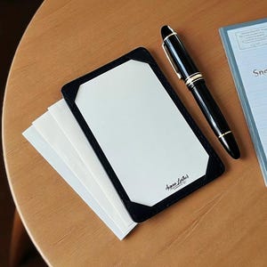 May include: A black leather notepad holder with white paper and the text "Argan Leather". A black fountain pen with gold accents rests beside the notepad. A small blue notebook is also visible.