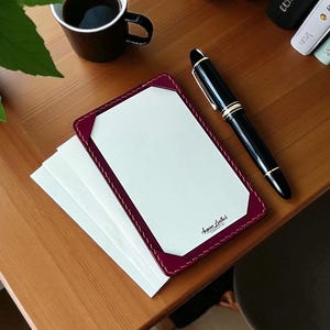 May include: A burgundy leather notepad with white paper, a black fountain pen, and a cup of coffee on a wooden surface. The notepad has a stitched border and the text "Aegan Leather". A stack of white paper is next to the notepad.