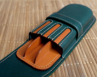 May include: A dark green leather case with a tan interior, designed to hold items. The case features white stitching and a secure flap closure. The interior has slots for organizing items. The case is made of high-quality leather.